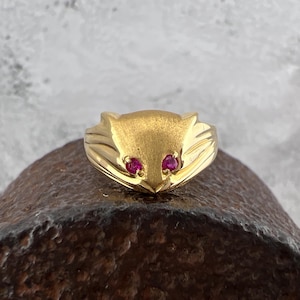 May include: A gold-toned ring featuring a stylized cat face design. The cat's eyes are represented by two small, round, red gemstones. The ring has a textured finish and is set against a neutral background.