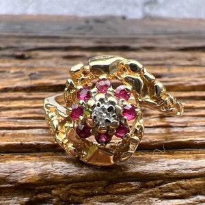 May include: A gold ring with a cluster of small, round, red gemstones and a central diamond. The ring has a textured, nugget-like design around the gemstones. The ring is set on a wooden surface.
