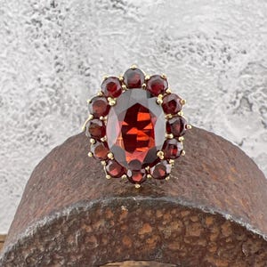 May include: A gold ring with a large oval-shaped red garnet surrounded by smaller red garnets.