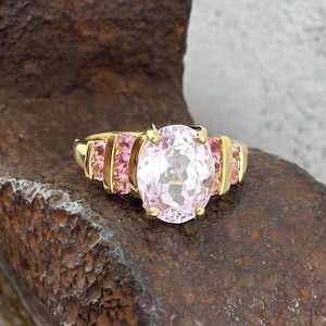 Könnte beinhalten: Ein goldener Ring mit einem großen, oval geschliffenen rosa Edelstein im Mittelpunkt. Der Ring ist mit kleineren rosa Edelsteinen auf jeder Seite verziert, die in einem goldenen Band gefasst sind. Der Ring wird auf einer strukturierten, braunen Oberfläche präsentiert.