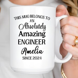 Könnte beinhalten: Weiße Keramiktasse mit schwarzem Text: „THIS MUG BELONGS TO AN Absolutely Amazing ENGINEER Amelia SINCE 2024“. Die Tasse hat einen großen Henkel und wird von einer Person gehalten.
