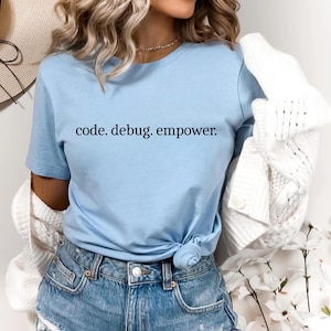 May include: Light blue t-shirt with the text "code. debug. empower." in black. The shirt is knotted at the waist and paired with denim shorts. A white cardigan is draped over the shoulders.