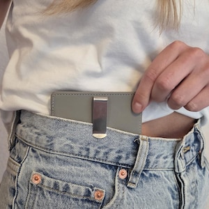 Puede incluir: Un monedero con clip gris con un clip de metal plateado es parcialmente visible en el bolsillo delantero de unos vaqueros azules. Los vaqueros tienen remaches color cobre y un diseño clásico.