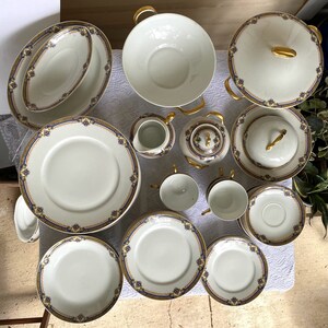 May include: A complete white porcelain dinnerware set with gold trim and blue floral accents. The set includes plates, bowls, cups, saucers, a teapot, a creamer, and a covered serving dish. The handles and accents are gold.