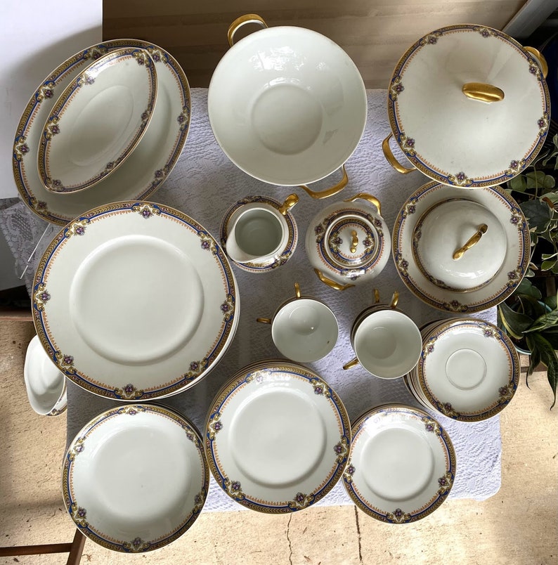 May include: A complete white porcelain dinnerware set with gold trim and blue floral accents. The set includes plates, bowls, cups, saucers, a teapot, a creamer, and a covered serving dish. The handles and accents are gold.