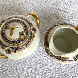 May include: A cream-colored tea set with gold accents. The set includes a lidded teapot and a creamer. Both pieces feature a blue and gold decorative border with floral details. The handles and lid finial are gold.