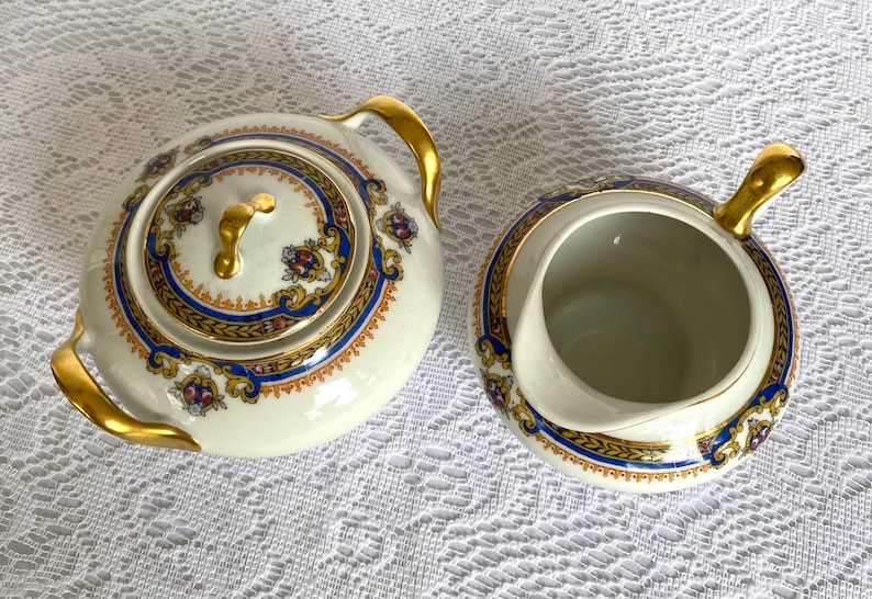 May include: A cream-colored tea set with gold accents. The set includes a lidded teapot and a creamer. Both pieces feature a blue and gold decorative border with floral details. The handles and lid finial are gold.