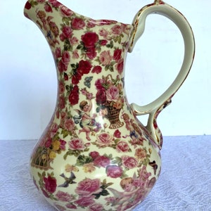 May include: A cream-colored ceramic pitcher with a floral pattern featuring pink and red roses, cherubs, and butterflies. The handle is gold-trimmed.
