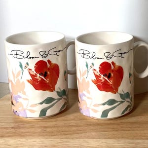 May include: Two white ceramic mugs with a floral design. The mugs have a black inscription that reads "Bloom & Grow".