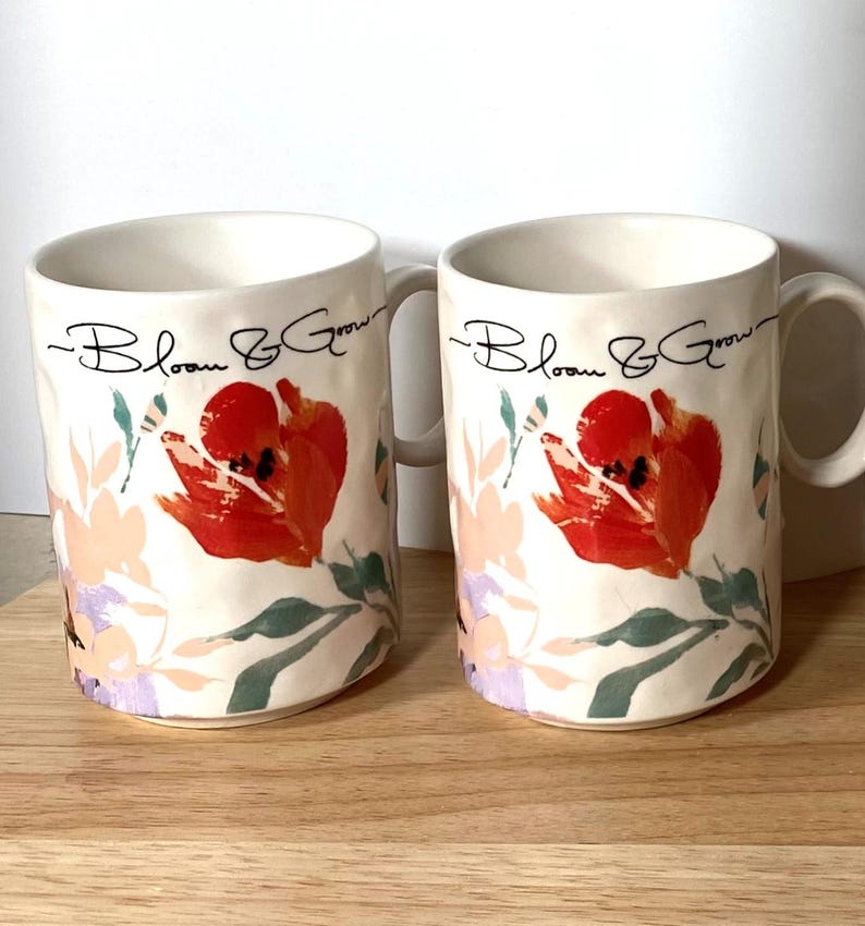 May include: Two white ceramic mugs with a floral design. The mugs have a black inscription that reads "Bloom & Grow".