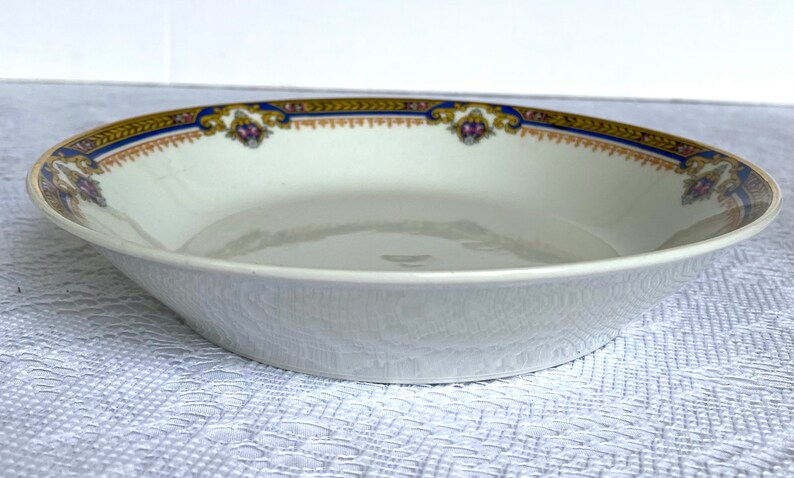 Puede incluir: Un taz&oacute;n de cer&aacute;mica blanca con un borde azul y dorado y un dise&ntilde;o floral. El taz&oacute;n est&aacute; vac&iacute;o y se encuentra sobre un mantel blanco.