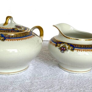 May include: A white porcelain sugar bowl with a lid and a creamer. Both pieces have gold handles and trim, with a blue and gold decorative band. The sugar bowl has a matching lid with a gold handle.