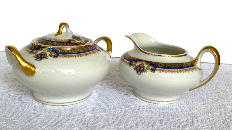 May include: A white porcelain sugar bowl with a lid and a creamer. Both pieces have gold handles and trim, with a blue and gold decorative band. The sugar bowl has a matching lid with a gold handle.