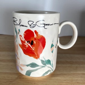 May include: White ceramic mug with a floral design featuring a red tulip and green leaves. The mug has black text that reads "Bloom & Grow".