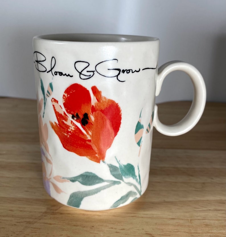 May include: White ceramic mug with a floral design featuring a red tulip and green leaves. The mug has black text that reads "Bloom & Grow".