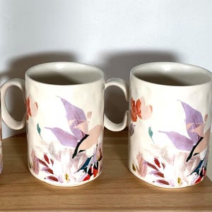 May include: Set of three white ceramic mugs with a floral design featuring pink, purple, and green accents. The mugs have a textured surface and a rounded handle.