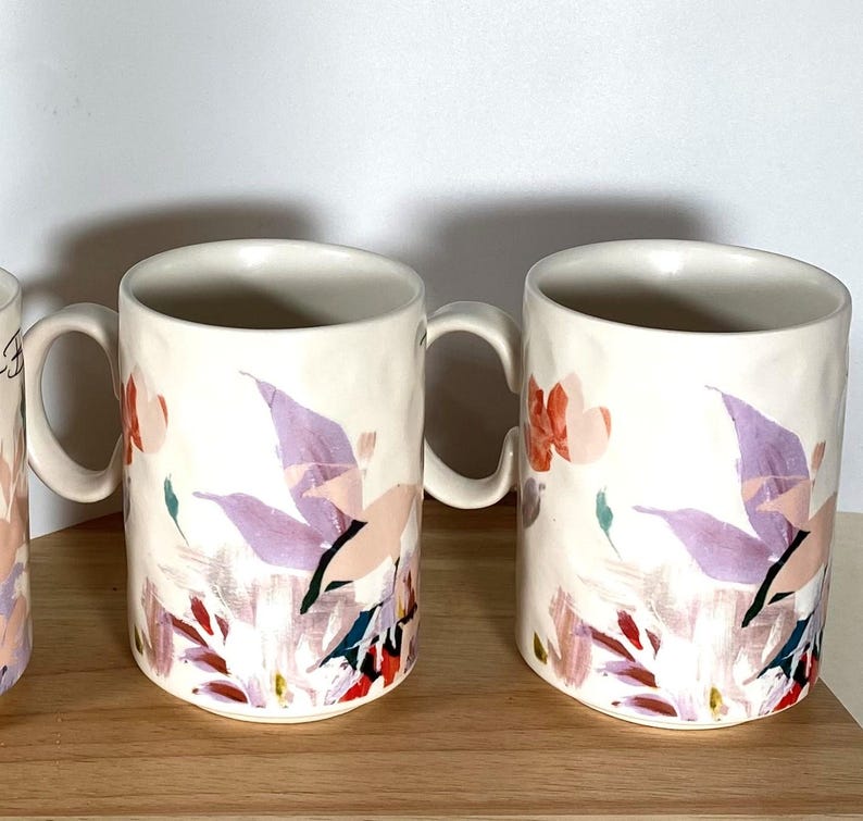 May include: Set of three white ceramic mugs with a floral design featuring pink, purple, and green accents. The mugs have a textured surface and a rounded handle.