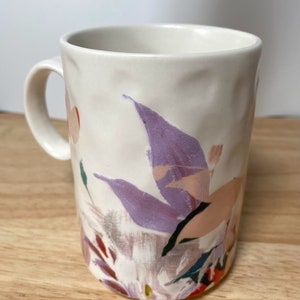May include: White ceramic mug with a floral design featuring purple, pink, and green abstract flowers. The mug has a textured surface and a handle.