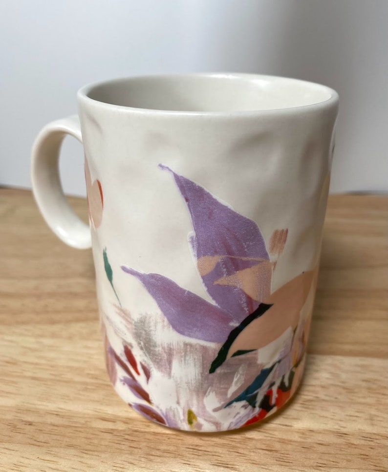 May include: White ceramic mug with a floral design featuring purple, pink, and green abstract flowers. The mug has a textured surface and a handle.