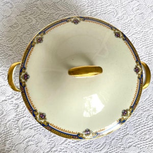 May include: Cream-colored, lidded serving dish with gold handles and accents. The rim features a blue and gold border with floral details. The lid has a gold handle. A decorative piece for serving food.