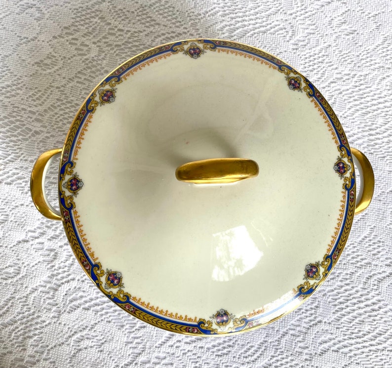 May include: Cream-colored, lidded serving dish with gold handles and accents. The rim features a blue and gold border with floral details. The lid has a gold handle. A decorative piece for serving food.