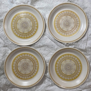 Puede incluir: Juego de cuatro platos de cena de cerámica blanca con un patrón geométrico dorado y marrón. Los platos tienen un borde elevado y una superficie ligeramente texturizada.