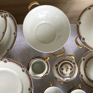 May include: A collection of white porcelain dinnerware with gold trim and blue and burgundy floral accents. The set includes serving bowls, plates, a teapot, and a creamer. The handles and lids have gold accents.