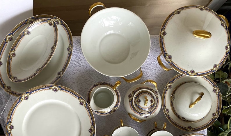 May include: A collection of white porcelain dinnerware with gold trim and blue and burgundy floral accents. The set includes serving bowls, plates, a teapot, and a creamer. The handles and lids have gold accents.