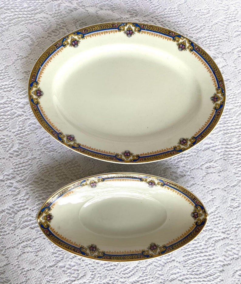 May include: Two oval serving platters. The larger platter is approximately 16 inches long, and the smaller one is about 9 inches long. Both have a cream-colored surface with a decorative border of gold, blue, and purple accents.