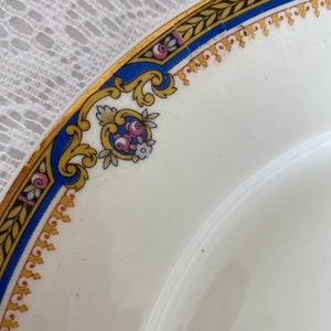 Puede incluir: Primer plano del borde de un plato de cer&aacute;mica blanca con un dise&ntilde;o floral dorado, azul y rosa. El plato tiene un borde dorado con un patr&oacute;n repetitivo de hojas y una banda azul con un patr&oacute;n repetitivo de peque&ntilde;os cuadrados.