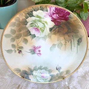 Puede incluir: Un tazón grande de porcelana blanca con un borde dorado. El tazón está decorado con un patrón floral de rosas rosas y blancas con hojas verdes.