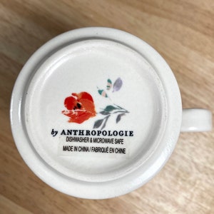 May include: White ceramic mug with a floral design featuring a red flower and green leaves. The mug has the text "by Anthropologie" and "Dishwasher & Microwave Safe" printed on the bottom. The text "Made in China / Fabriqu&eacute; en Chine" is also printed on the bottom.