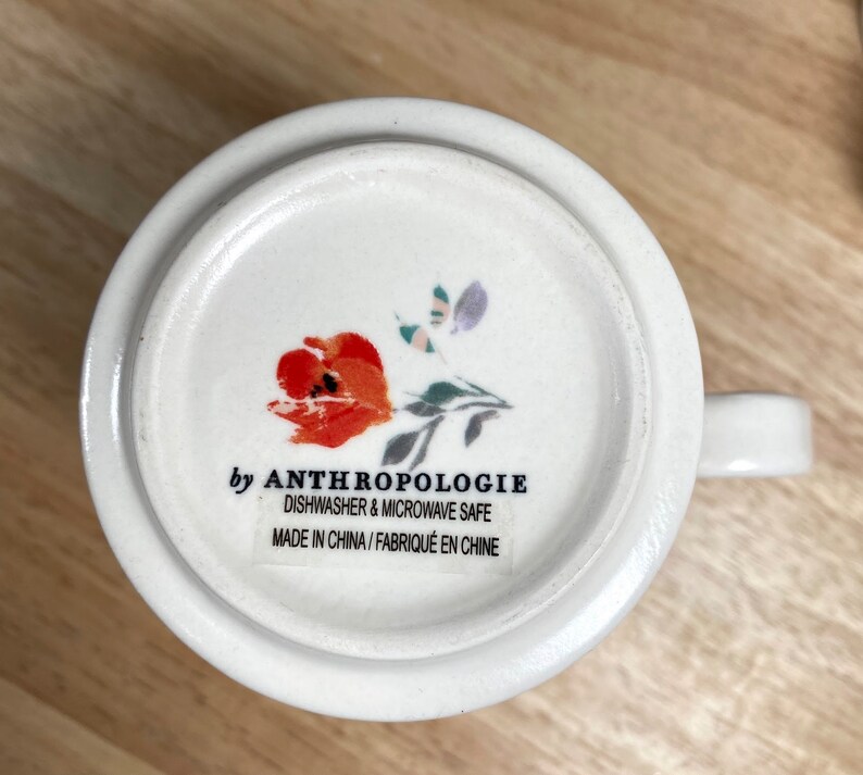 May include: White ceramic mug with a floral design featuring a red flower and green leaves. The mug has the text "by Anthropologie" and "Dishwasher & Microwave Safe" printed on the bottom. The text "Made in China / Fabriqu&eacute; en Chine" is also printed on the bottom.