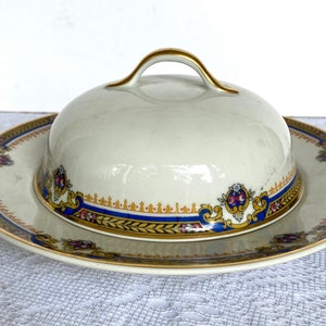 May include: Cream-colored ceramic butter dish with a matching lid. The dish and lid feature a decorative border with blue, gold, and red floral accents. The lid has a gold handle.
