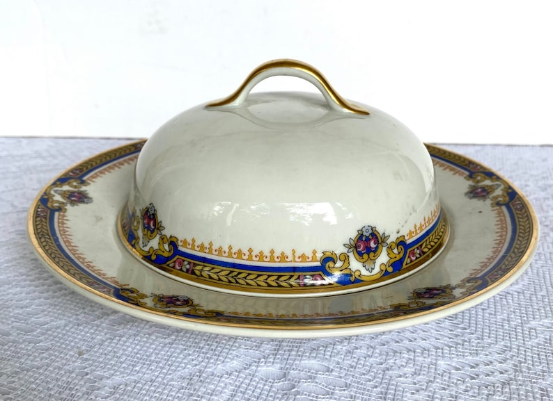 May include: Cream-colored ceramic butter dish with a matching lid. The dish and lid feature a decorative border with blue, gold, and red floral accents. The lid has a gold handle.