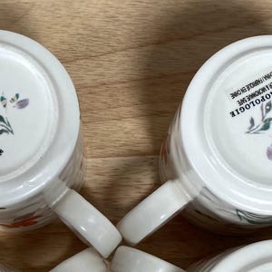 May include: Four white ceramic mugs with a floral pattern. The mugs feature a red poppy design and the text "by Anthropologie" on the bottom.
