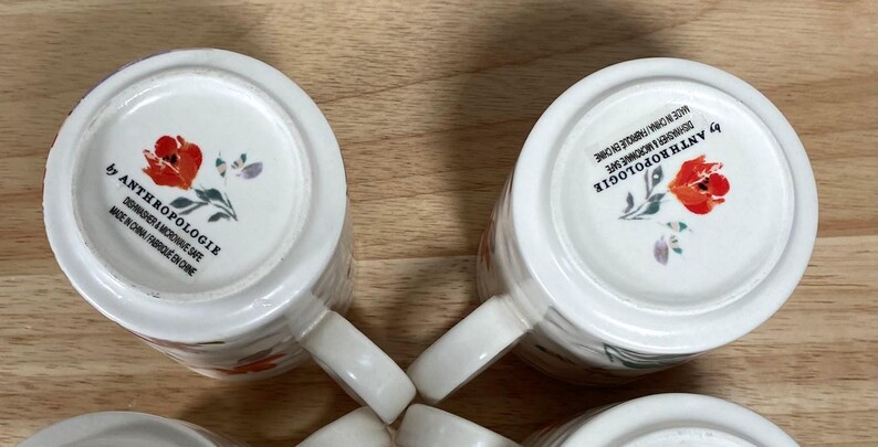May include: Four white ceramic mugs with a floral pattern. The mugs feature a red poppy design and the text "by Anthropologie" on the bottom.