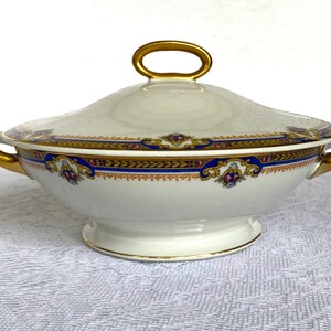 May include: A white porcelain tureen with a lid, featuring gold handles and a decorative band of blue, gold, and maroon accents. The tureen is designed for serving soups or stews.