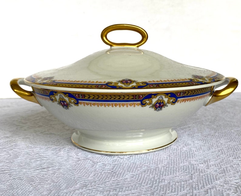 May include: A white porcelain tureen with a lid, featuring gold handles and a decorative band of blue, gold, and maroon accents. The tureen is designed for serving soups or stews.