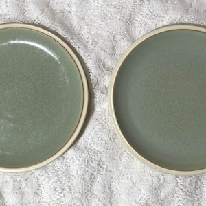May include: Two green ceramic plates with a white rim. The plates are on a white lace tablecloth.