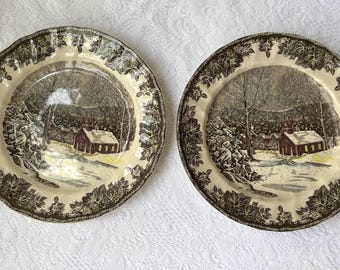 May include: Two brown and white ceramic dinner plates with a winter scene of a red schoolhouse in a snowy forest. The plates have a scalloped edge and a gold trim.