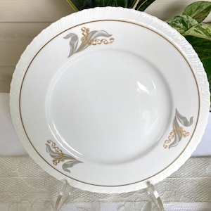May include: A white porcelain plate with a gold rim and a delicate floral design featuring gray lily of the valley flowers and leaves.
