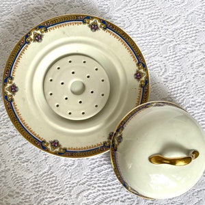 May include: A cream-colored, antique porcelain serving dish with a lid. The dish has a decorative gold, blue, and purple border. The lid has a gold handle. The dish has a center piece with multiple holes.