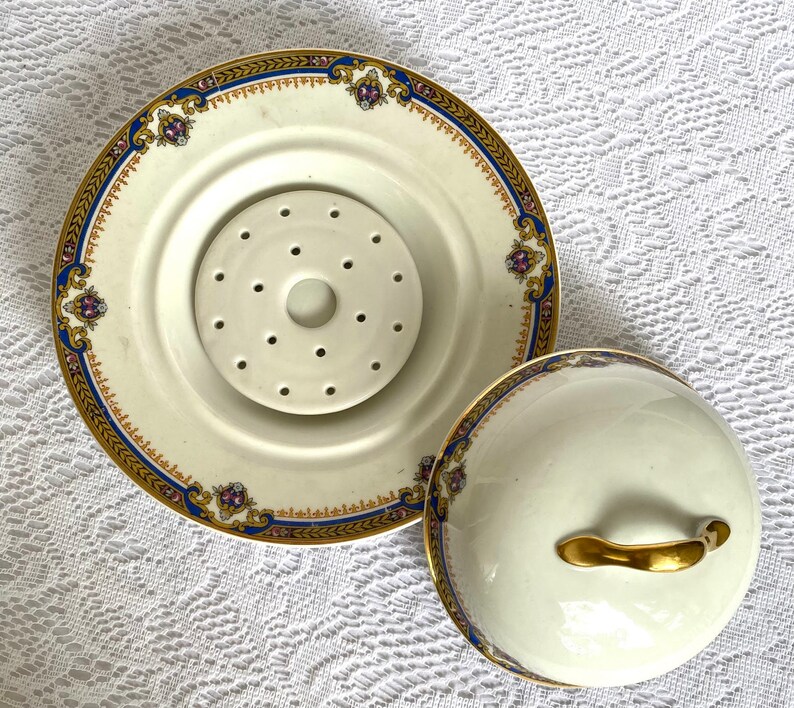 May include: A cream-colored, antique porcelain serving dish with a lid. The dish has a decorative gold, blue, and purple border. The lid has a gold handle. The dish has a center piece with multiple holes.