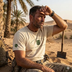 Puede incluir: Un hombre en un entorno desértico lleva una camiseta beige con el texto "INGREDIOR - ADEPTU - EX" y "52ND INFANTRY DIVISION". La camiseta tiene un emblema verde. Lleva un reloj negro y pantalones de camuflaje.