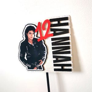 Puede incluir: Un adorno para pastel con un recorte de Michael Jackson en chaqueta de cuero negra, el número 12 en rojo brillante y el nombre HANNAH en letras negras con purpurina sobre un fondo blanco brillante, montado en un palo negro fino.