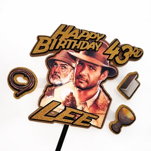 May include: A cake topper featuring two men, one wearing a fedora. The words "HAPPY BIRTHDAY 43rd LEE" are in gold glitter. Additional themed cutouts include a whip, a map, and a chalice.