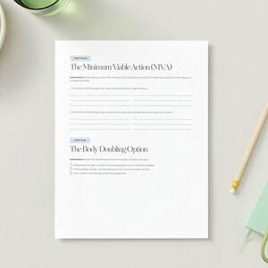 Pode incluir: Uma folha de papel branca com texto, um l&aacute;pis, um pequeno caderno verde e um cacho de uvas verdes numa tigela branca. O papel tem o t&iacute;tulo "The Minimum Viable Action (MVA)" e "The Body Doubling Option".