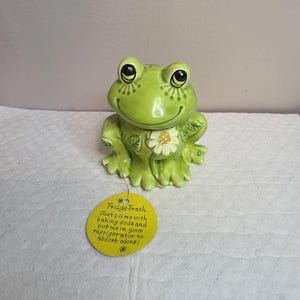 May include: A light green ceramic frog figurine with a white daisy accent on its chest. A round yellow tag is attached, featuring the text: Fridge-Fresh. Just fill me with baking soda and put me in your refrigerator to absorb odours!