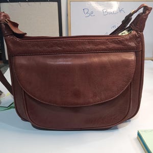 May include: A brown leather shoulder bag with a flap closure and a zippered top. The bag has a single strap and a small pocket on the front.